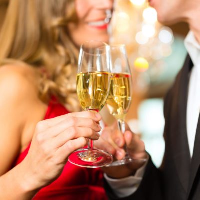 Eine blonde Dame stößt mit einem Callboy bei einem Sektglas an. Beide sind festlich gekleidet