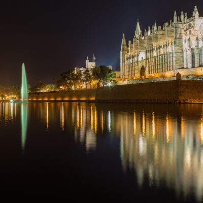 In this captivating scene, the majestic cathedral stands as a true masterpiece of Gothic architecture. Its intricate details are intricately highlighted by strategic lighting, casting a warm glow that accentuates its grandeur against the night sky. The reflection on the tranquil water doubles its beauty, creating a mesmerizing mirror image that captures the eye. To the left, a tall fountain catches attention with its cascading waters glistening under soft lights. The gentle sound of flowing water adds to the serene ambiance of the setting. Surrounding buildings are tastefully illuminated, adding depth and context to this picturesque view without overpowering the cathedrals commanding presence. Together, these elements form a harmonious blend of history and modernity—a scene steeped in elegance and allure akin to an enchanting evening stroll along Mallorcas vibrant coastlines.