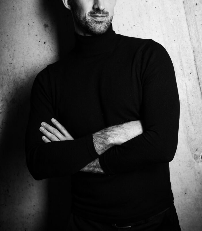 Die Schwarz-Weiß-Ästhetik des Fotos von Callboy Ryan verleiht ihm eine zeitlose und geheimnisvolle Qualität, die durch seine Kleidungswahl – einen schwarzen Rollkragenpullover – noch verstärkt wird. Die Betonwand im Hintergrund sorgt für einen starken Kontrast, der ihn in den Fokus rückt und gleichzeitig die minimalistische Erscheinung des Fotos verstärkt. Seine verschränkten Arme strahlen Selbstvertrauen aus, und die teilweise Verdeckung seiner Identität durch die fotografische Betonung seines Mundes und Kinns führt ein faszinierendes Element ein. Diese Komposition lädt den Betrachter ein, über seine Geschichte nachzudenken und gleichzeitig das künstlerische Gleichgewicht zwischen Licht, Schatten und Textur im Bild zu würdigen.