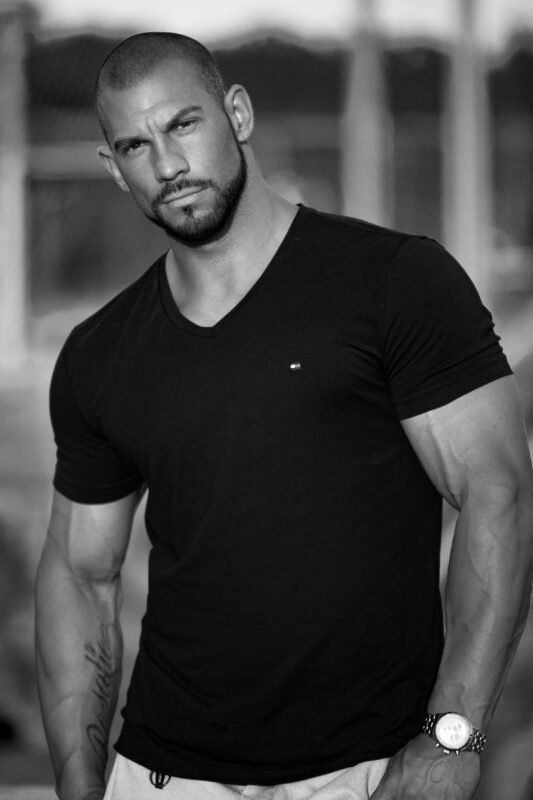 Das Schwarz-Weiß-Bild von Callboy Alex strahlt eine zeitlose und geheimnisvolle Aura aus und unterstreicht seine imposante Präsenz. Das taillierte schwarze T-Shirt betont seinen muskulösen Körperbau, während die Uhr seinem Ensemble einen subtilen Hauch von Raffinesse verleiht. Sein Bart trägt zu dem rauen Charme bei, der seinen ernsten Gesichtsausdruck ergänzt. Der unscharfe Hintergrund mit Bäumen und einem Zaun verstärkt den Fokus auf ihn und schafft ein intimes und dennoch kraftvolles Porträt, das die Neugier auf den Mann hinter dem rätselhaften Äußeren weckt.