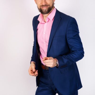 This image description suggests a charismatic and stylish individual, exuding confidence and approachability. The combination of the blue suit and pink shirt creates a striking contrast, adding to the visual appeal. His bearded appearance and ponytail give off a modern, relaxed vibe while maintaining an air of professionalism through his attire. The smile and gesture of pointing forward can be interpreted as engaging or inviting, suggesting an openness to interaction or communication with viewers.