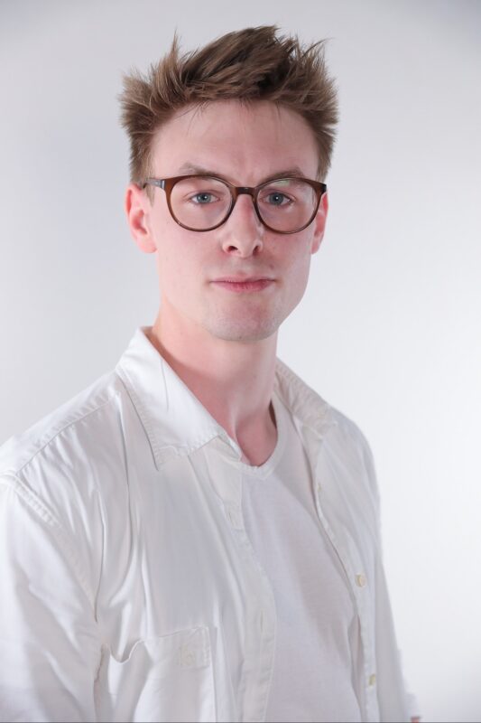 It sounds like you are describing either a reference image for art or perhaps requesting a descriptive summary for a scene or character. Here’s an expanded characterization based on your description: A young man stands against a simple, light-colored backdrop. His short brown hair is tousled, giving him an effortlessly casual look. He wears glasses that complement his thoughtful appearance and dresses simply—a crisp white button-up shirt layered over a plain white t-shirt. His expression is neutral as he looks straight into the camera, neither smiling nor frowning, creating an open and approachable yet reserved demeanor. If you meant this as a prompt for generating or searching for such an image—feel free to clarify if you need it in another format (e.g., detailed for artists, AI image prompts, etc.).