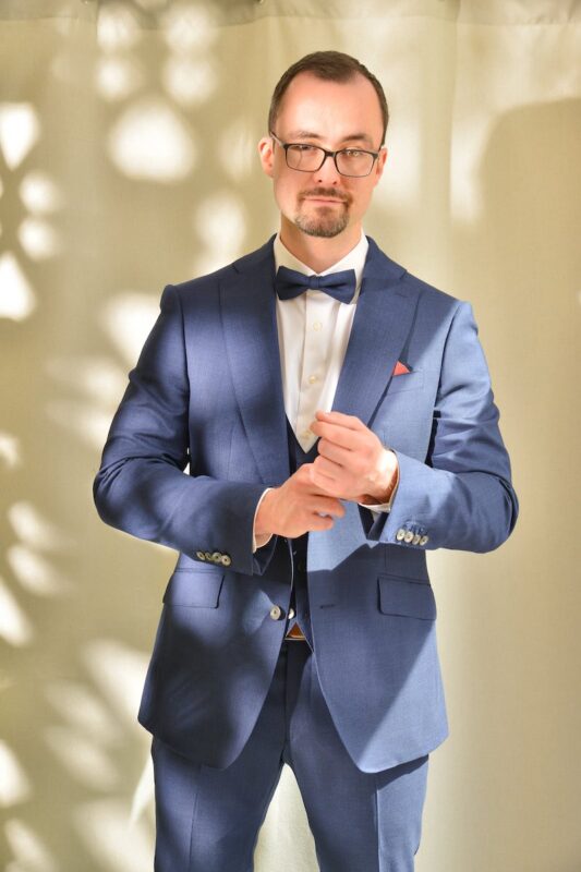 Callboy Alexander steht vor einem sanft beleuchteten, gesprenkelten Hintergrund. Er trägt eine elegante Brille und einen maßgeschneiderten blauen Anzug, komplettiert durch ein frisches weißes Hemd und eine stilvolle Fliege. Mit einer Hand rückt er sorgfältig seine Manschettenknöpfe zurecht und strahlt ruhiges Selbstvertrauen aus. Das Sonnenlicht fällt sanft durch und betont sowohl die klaren Linien seiner Kleidung als auch die gelassene Sicherheit in seinem Gesichtsausdruck.