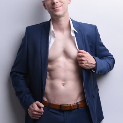 A possible description for your scene could be: Dennis stands confidently against a plain light background, dressed in a sharp blue suit. His white shirt is unbuttoned to reveal a toned chest and abs, adding an air of casualness to his formal attire. With a friendly, inviting smile directed at the camera, Dennis exudes both charm and confidence.